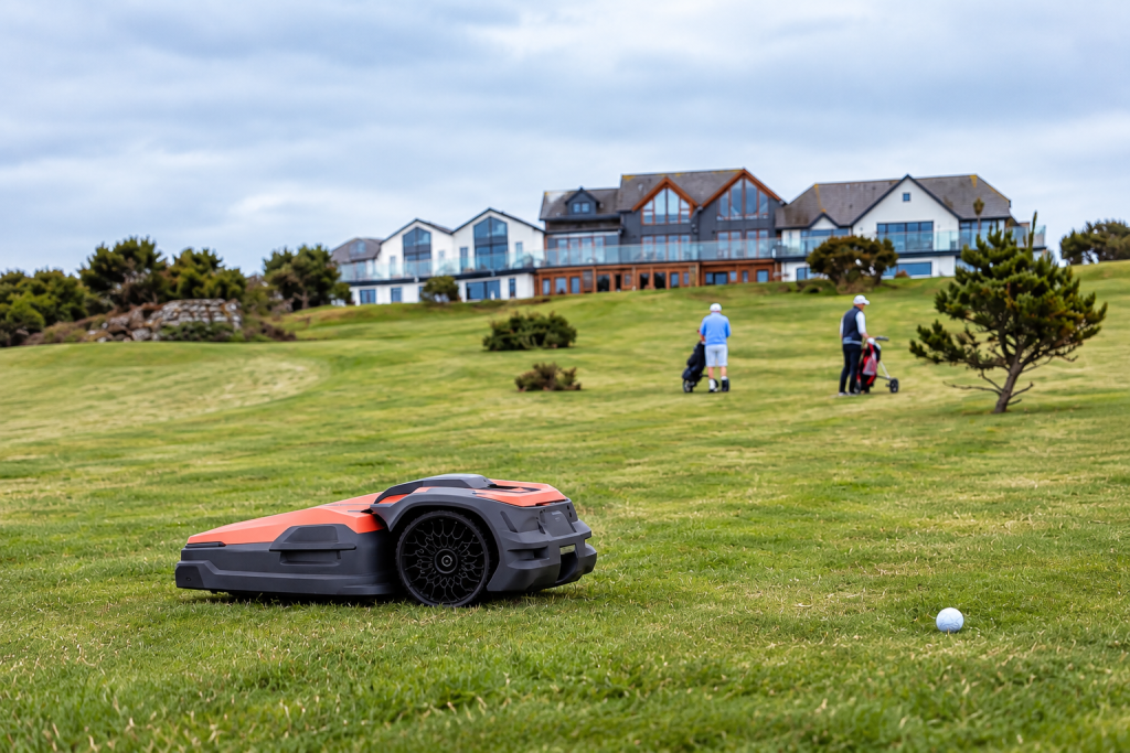 Autonomous robotic mower operating at The Point at Polzeath in Cornwall with golfers and the clubhouse in view